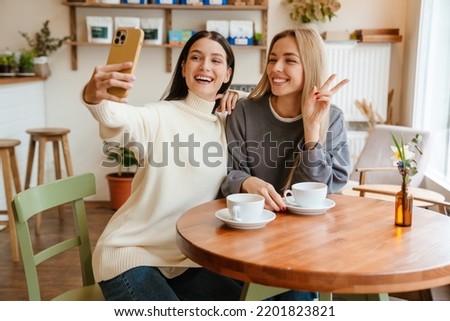 Two young women gesturing while taking selfie photo on mobile phone at cafe indoors