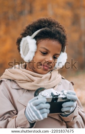 Cute African-American girl in fur headphones takes pictures with a camera in an autumn park.Diversity,autumn concept.