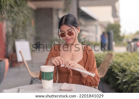A beautiful young lady wearing frameless sunglasses attempts to open the disposable straw for her drink.