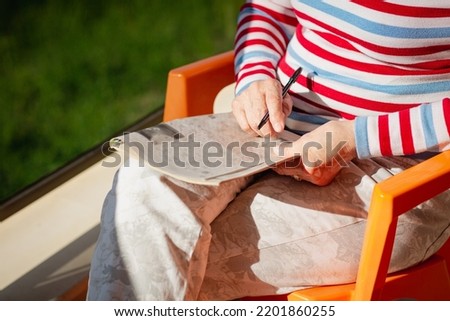 Elderly unrecognizable woman in glasses sitting on a balcony near the sea and looking at magazine, solving a crossword Royalty-Free Stock Photo #2201860255