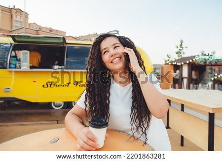 A photo of a young smiling happy woman who sits at the food court of a shopping center at a table and works at a computer laptop, talks on a mobile phone. Freelance concept