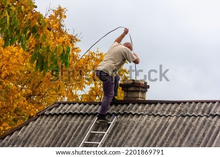 a male chimney sweep cleans the chimney from soot on the roof of a village house, preparation for the heating season Royalty-Free Stock Photo #2201869791