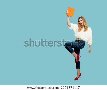 Happy stylish fashion student girl in jeans and red high heel shoes holds an orange textbook and rejoices in her success. Portrait of a college student on a blue background