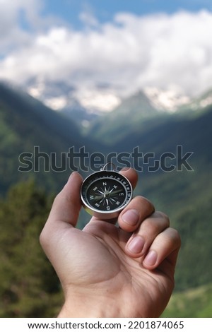 Magnetic compass in the palm of a male hand against the backdrop of a mountain range in the clouds in the summer outdoors, travel, first-person view Royalty-Free Stock Photo #2201874065