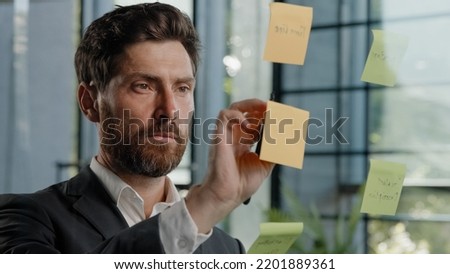 Close up busy focused smart male manager worker CEO boss planning work strategy pensive thoughtful bearded Caucasian businessman writing ideas on sticky notes glues on glass wall in office organized