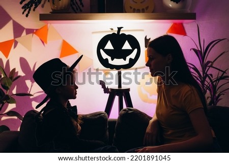 Silhouette of small kid daughter and young mom telling spooky stories sitting on comfy couch in room decorated for bats and pumpkins. Mother and child girl have fun at home. Happy Halloween concept