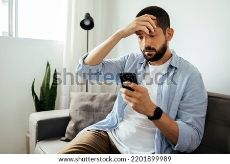 Sad man checking smartphone sitting on a sofa at home Royalty-Free Stock Photo #2201899989