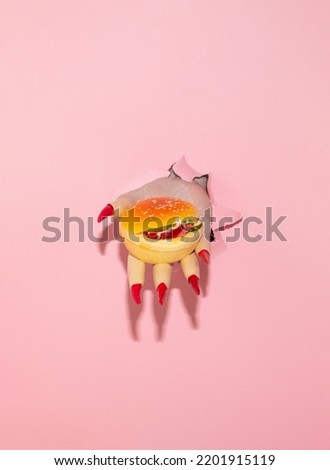 Witch hand holding hamburger. Pastel pink torn paper. Halloween surreal spooky idea. Minimal food concept.