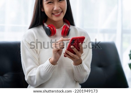 A beautiful Asian woman is wearing headphones and choosing a song from her smartphone. and relax on the sofa at home on vacation