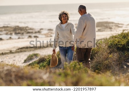 Happy elderly woman pulling her husband by the hand while going to the beach for a picnic. Cheerful senior couple taking a seaside holiday after retirement.