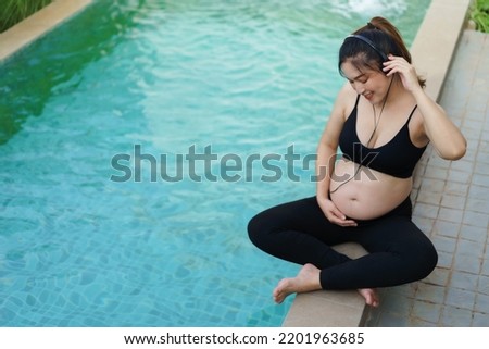 young pregnant woman sitting on edge of swimming pool while listening to music with headphone