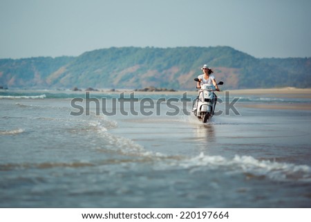 Wide ocean shore with small waves on which rides a young woman in white clothes on his scooter.
