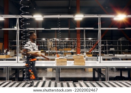 African man working at the factory, walking along the production line with box, delivering order. He is wearing uniform. He is moving in motion blur, factory interior staying still