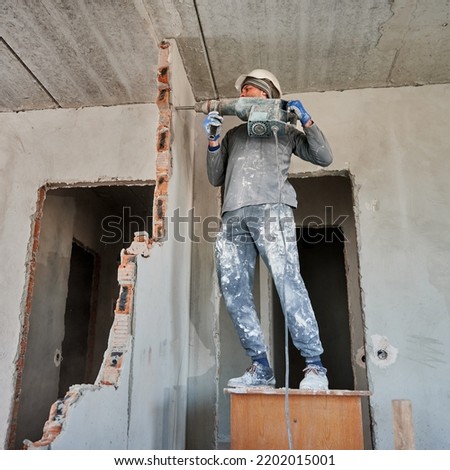 Full length of man in workwear drilling wall with hammer drill. Male worker using drill breaker while destroying wall in apartment under renovation. Demolition work and home renovation concept. Royalty-Free Stock Photo #2202015001