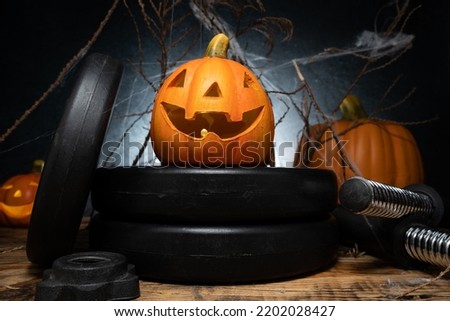 Small ceramic Halloween pumpkin on a dumbbells barbell weight plates. Healthy gym fitness lifestyle autumn or fall composition with decorative Jack-o'-lantern spooky laughing, scary head.