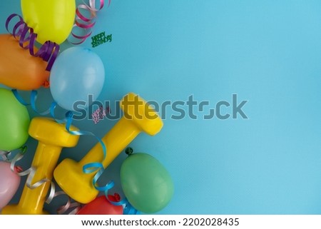 Two yellow dumbbells, colorful balloons and ribbons. Gym exercise equipment as a gift for birthday party. Healthy fitness lifestyle flat lay composition concept with copy space on blue background.