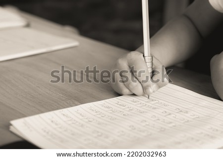 The kid's hand is practicing and learning to write English cursive handwriting sentences in a notebook with a pencil. Kindergarten writing skills. Sepia, monochrome, black and white color background. Royalty-Free Stock Photo #2202032963