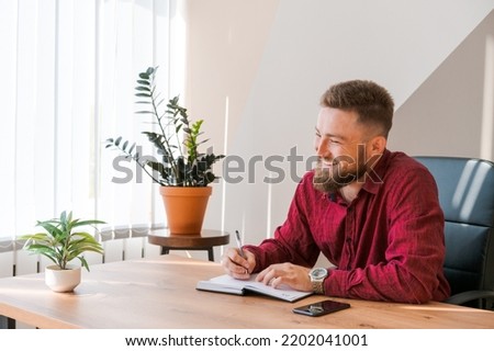 Portrait handsome smiling man with beard, working in office on some project, he sits at the table, writes ideas in his notepad or diary. Thinking about a successful business deal Royalty-Free Stock Photo #2202041001