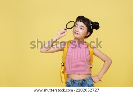 Asian child schoolgirl stood in casual wear looking at camera through magnifier while standing bright yellow background. Royalty-Free Stock Photo #2202052727