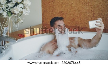 Cheerful young man with foam on beard is taking selfie using smartphone in hot tub in modern spa salon. He is laughing, gesturing, posing and looking at camera.