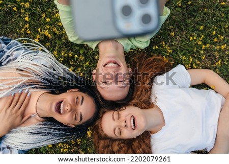Top view of a group of cheerful bloggers shooting selfie video reading funny comments on website while lying on a grassy lawn. Sunny day and laugh. Emotions and