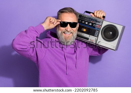 Photo of old handsome good mood man wear purple hoodie fingers touch sunglass hold boombox on shoulder isolated on violet color background