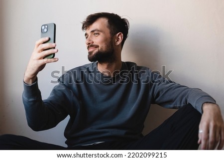 Handsome brunette man sitting on floor holding mobile phone and make selfie, video call or use smartphone like mirror, look at himself smiling, joy of his look against on white wall background at home