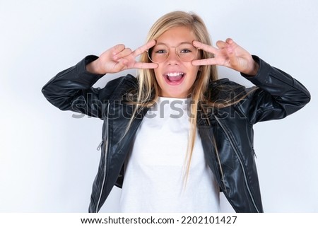 Cheerful positive beautiful caucasian blonde little girl wearing biker jacket and glasses over white background shows v-sign near eyes open mouth