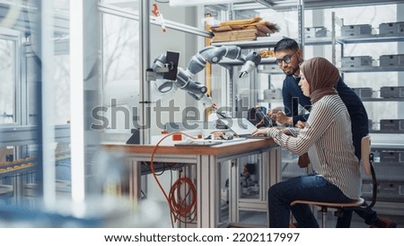 Black Male Engineer Raises Robotic Arm with Controller While Female Developer in Hijab Writes Code on Laptop. People Create Innovative Application. High Tech and Robotics Industries Concept.