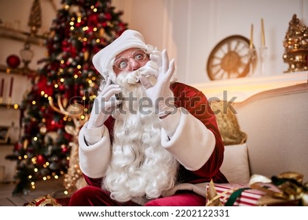 Santa Claus sitting on couch and talking on mobile phone near the fireplace and christmas tree with gifts. New year and Merry Christmas , happy holidays concept