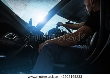 Caucasian Middle Aged Rich Man Sitting on a Driver's Seat in His Super Car Keeping Right Hand on the Steering Wheel and Getting Ready to Leave the Underground Garage. Royalty-Free Stock Photo #2202145233
