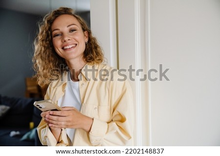 Young beatiful attractive smiling happy curly woman holding phone and looking at camera while standing at home Royalty-Free Stock Photo #2202148837