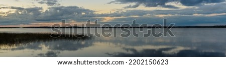 widescreen panoramic view of a large lake with reeds and a wooded distant shore in a foggy haze under a cloudy sky reflected in the water. serene morning twilight