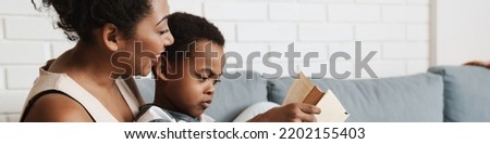 Black pleased mother and son reading book while sitting on sofa at home