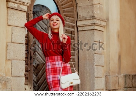 Fashionable happy smiling blonde woman wearing stylish red beret,  cashmere sweater, checkered skirt, holding trendy white bag, posing in street of European city. Copy, empty space for text 