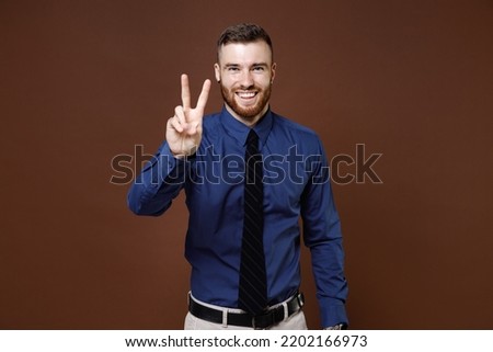 Cheerful smiling bearded young business man wearing blue shirt tie showing victory sign looking camera isolated on brown colour background studio portrait. Achievement career wealth business concept