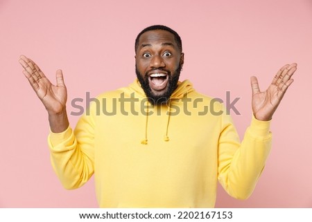 Shocked surprised young man 20s wearing casual basic yellow streetwear hoodie standing keeping mouth open spreading hands isolated on plain light pastel pink color background studio portrait
