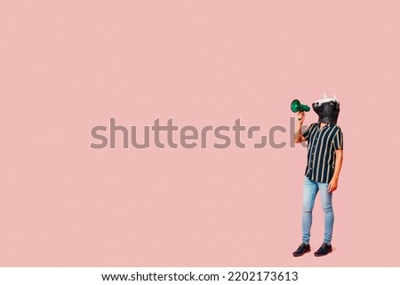 a standing young man wears a cow mask and speaks into a green megaphone, on a pink background with some blank space on the left