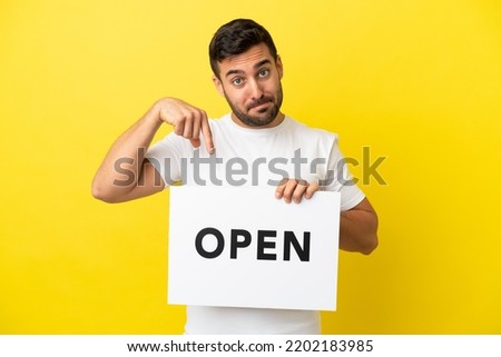 Young handsome caucasian man isolated on yellow background holding a placard with text OPEN and  pointing it