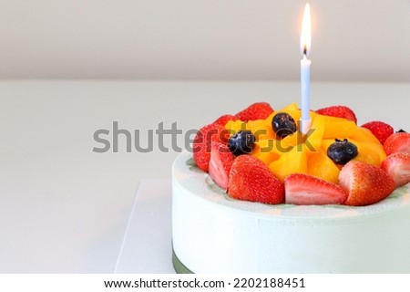 Delicious birthday cake, fruit cake (with fresh strawberries, blueberries and mango) with light blue candle lit with flame, on right side of frame, copy space on left