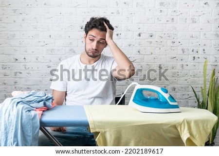 Tired and upset man sitting behind ironing board with many clothes on it. Household and exhausting ironing concept