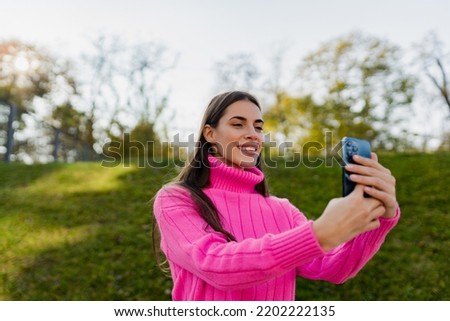 portrait of candid attractive young smiling woman in pink sweater walking in green park using phone happy mood, fashion style trend taking selfie pictures photo on smartphone camera
