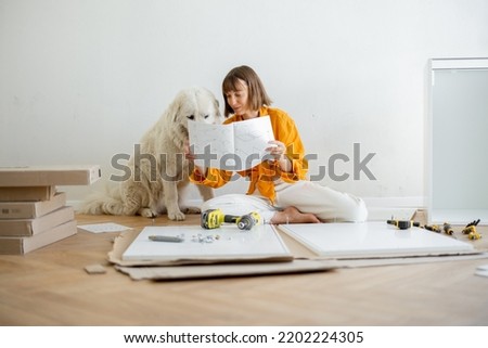 Young woman assembles furniture by herself, sitting with dog and reading manual at new apartment. DIY and house improvement concept Royalty-Free Stock Photo #2202224305