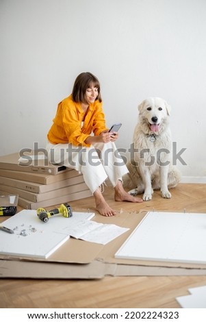 Young woman sits with her cute dog, using phone during repairing at new apartment. Assembling furniture by herself. DIY concept Royalty-Free Stock Photo #2202224323