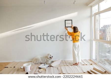 Young woman hanging picture frame in room, decorating her newly renovated apartment, stands with her dog in white room