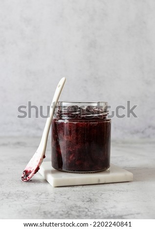 A glass jar with blackberry jam on a white marble stand. A white ceramic spoon next to the jar. Light background Royalty-Free Stock Photo #2202240841