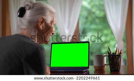 Over shoulder view of smiling senior elderly woman making video call on laptop green screen talking and listening during video chat with friends or colleagues.