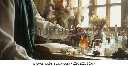 A close-up of a perfumer at his desk looking for a new fragrance. Vintage cinematic perfumery concept. Sunny day in the working workshop. Lots of ingredients, glass flasks. Depth of field Royalty-Free Stock Photo #2202251167