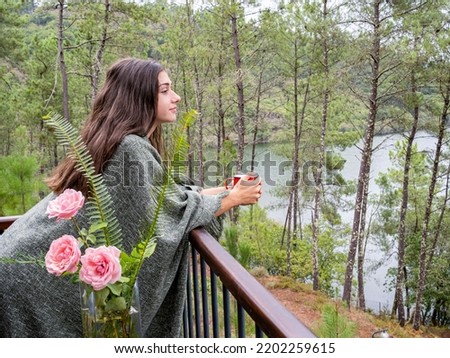 Portrait image of a beautiful Caucasian woman holding and drinking hot coffee , standing on the balcony and looking at the lake and green nature.