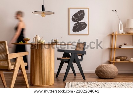 Warm and cozy interior of living room space with mock up poster frame, round table, chairs, pedant lamp, rattan chairs and walking young woman. Minimalist home decor. Template. Royalty-Free Stock Photo #2202284041